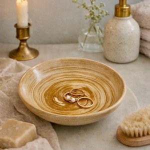May include: A decorative ceramic bowl with a swirling pattern holds several gold rings with pearls. A lit candle in a brass holder, a soap bar, and a soap dispenser are also visible, suggesting a bathroom or vanity setting.