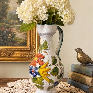 May include: A white ceramic pitcher vase with a speckled finish, featuring a hand-painted floral pattern in orange, yellow, blue, and green. The vase has a flared rim, a curved handle, and a pedestal base, currently holding a bouquet of white flowers.