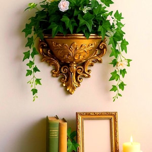 May include: A gold-colored wall-mounted planter filled with green ivy and small pink flowers. Below, a gold framed picture, two books, and a lit candle in a white holder sit on a wooden surface.