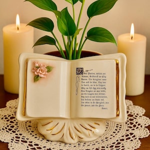 May include: A white ceramic book-shaped planter with gold trim, holding a green plant. The open book displays text and a pink rose. Two lit candles flank the planter, set on a lace doily.