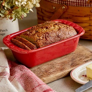 May include: A freshly baked loaf of bread in a bright red rectangular baking dish. Sliced bread is visible. The dish sits on a wooden cutting board, with a butter dish and knife in the foreground. A picnic basket and flowers are in the background.