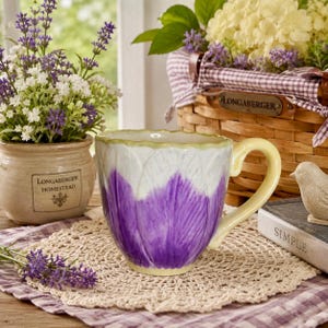 May include: A ceramic mug featuring a sculpted petal design with a white upper section and a deep purple lower section. The mug has a pale yellow handle and a scalloped rim, showcasing a textured surface that mimics the appearance of a flower bloom.