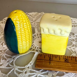 May include: A pair of ceramic salt and pepper shakers. One is shaped like a yellow and green ear of corn, the other like a yellow block with a white top. A wooden ruler is next to the shakers.