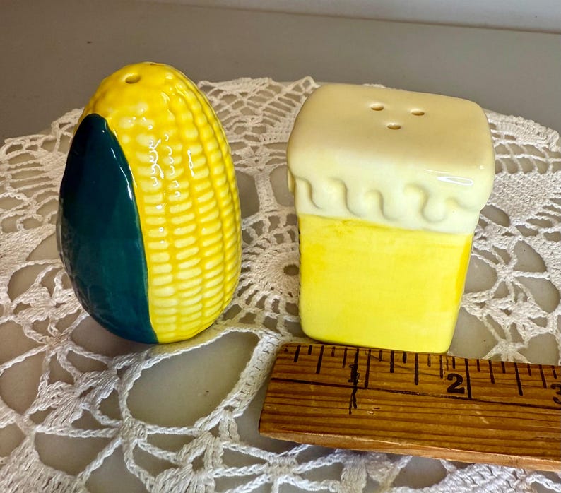 May include: A pair of ceramic salt and pepper shakers. One is shaped like a yellow and green ear of corn, the other like a yellow block with a white top. A wooden ruler is next to the shakers.
