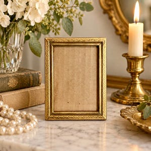 May include: A rectangular picture frame with a metallic gold finish and an embossed repeating geometric pattern along the border. The frame features a simple inner edge and a plain, light brown cardboard insert centered within the frame opening.