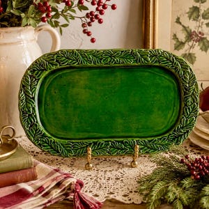 May include: An oval ceramic serving tray with a glossy deep green finish and a raised relief border featuring detailed oak leaves and acorns. The tray is displayed on a small gold-toned metal stand, showcasing its smooth center and textured rim.