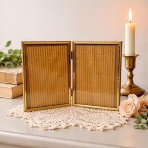 May include: A gold-toned, hinged double picture frame with a textured cardboard backing. The frame sits on a cream-colored lace doily, with a lit candle in a brass holder and a few roses in the background. The frame is empty.