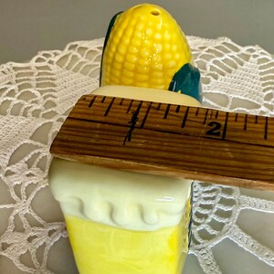May include: A ceramic salt shaker shaped like an ear of corn, with a yellow and green design. The shaker is approximately 2 inches tall. The top has a series of holes for dispensing salt. A charming kitchen accessory.