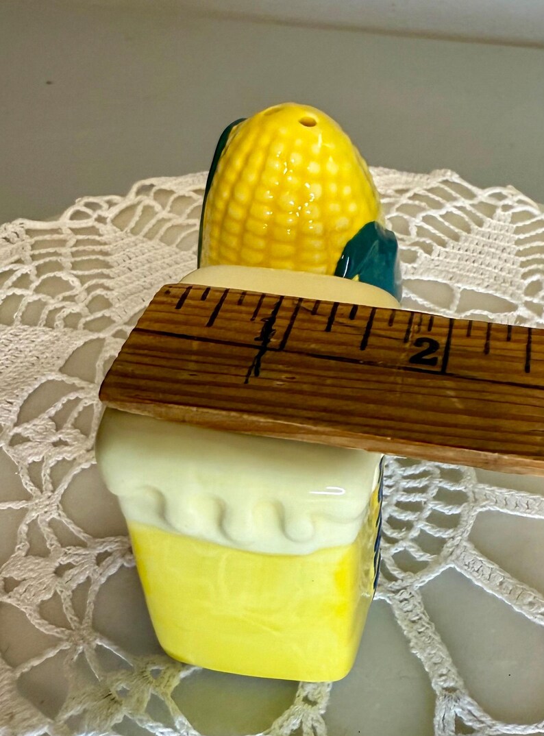 May include: A ceramic salt shaker shaped like an ear of corn, with a yellow and green design. The shaker is approximately 2 inches tall. The top has a series of holes for dispensing salt. A charming kitchen accessory.