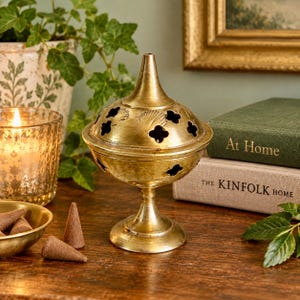 May include: A brass incense burner with a pointed lid and cut-out clover-shaped ventilation holes. The burner features a pedestal base and a metallic finish. It is accompanied by three small, cone-shaped incense pieces in a matching brass bowl.