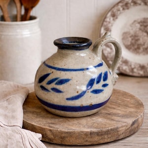 May include: A small, round ceramic pitcher with a handle, featuring a beige base and dark blue accents. The design includes a blue band and leaf-like patterns. It sits on a wooden cutting board, suggesting a rustic, farmhouse aesthetic.