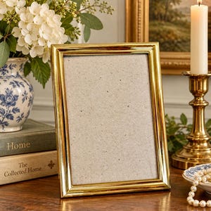 May include: A rectangular picture frame with a polished gold-tone metal finish and a simple beveled edge design. The frame is oriented vertically and contains a plain, light-colored speckled backing board, ready for a photograph or artwork display.