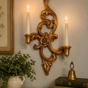 May include: A gold-colored wall sconce featuring an ornate, swirling scrollwork design with a central floral motif. The sconce holds two white taper candles, one on each side, which are lit and emitting a soft, warm glow against the wall.