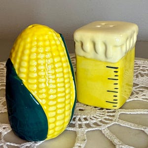 May include: A pair of ceramic salt and pepper shakers. One is shaped like an ear of corn, with yellow kernels and green husks. The other is a yellow cube with a white, frothy top and black measurement markings.