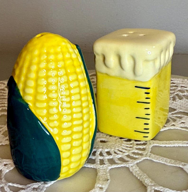 May include: A pair of ceramic salt and pepper shakers. One is shaped like an ear of corn, with yellow kernels and green husks. The other is a yellow cube with a white, frothy top and black measurement markings.