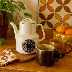 May include: A cream-colored ceramic teapot with a brown sunburst design, a matching lid, and two brown mugs sit on a wooden surface. A bowl of oranges and a floral patterned napkin are also present. The scene evokes a retro kitchen aesthetic.