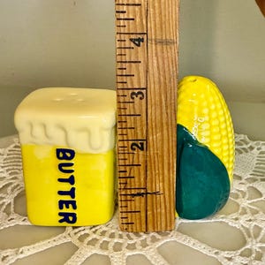 May include: A yellow ceramic butter-shaped salt shaker with the word "BUTTER" in blue. Next to it is a corn-shaped pepper shaker. The salt shaker is approximately 2.5 inches tall.