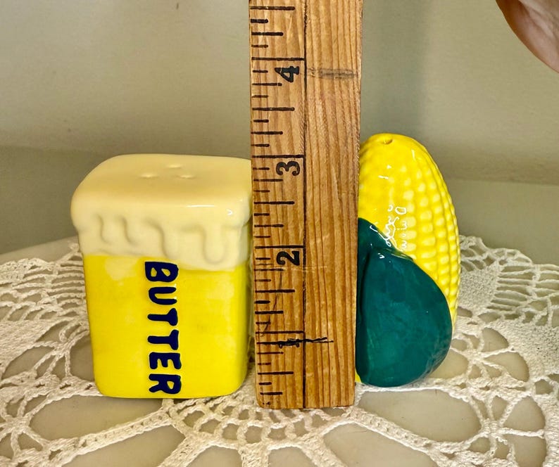 May include: A yellow ceramic butter-shaped salt shaker with the word "BUTTER" in blue. Next to it is a corn-shaped pepper shaker. The salt shaker is approximately 2.5 inches tall.