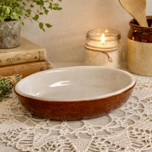 May include: Oval brown and white ceramic baking dish on a white lace doily. The dish is surrounded by a candle, wooden spoons in a ceramic jar, and a small potted plant. A stack of old books is in the background.