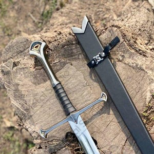 May include: A silver and black sword with a decorative hilt and crossguard, lying on a tree stump. The sword has a long, straight blade and is next to a black sheath. The hilt features gold and silver accents.