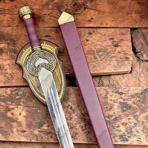 May include: A decorative sword with a silver blade, a burgundy handle, and a gold-colored hilt. The sword is displayed with a matching burgundy sheath and a decorative wooden plaque with gold accents. The words "Myth And Magic Studio" are visible.