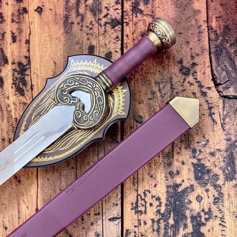 May include: A decorative sword with a silver blade, a burgundy handle, and a gold-colored hilt. The sword rests on a brown shield with gold filigree. A burgundy sheath with gold accents is also present.