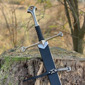 May include: A long, silver and black sword with a black sheath. The sword has a silver crossguard and a black handle. The sheath is secured with a black leather strap. The sword is resting on a tree stump.