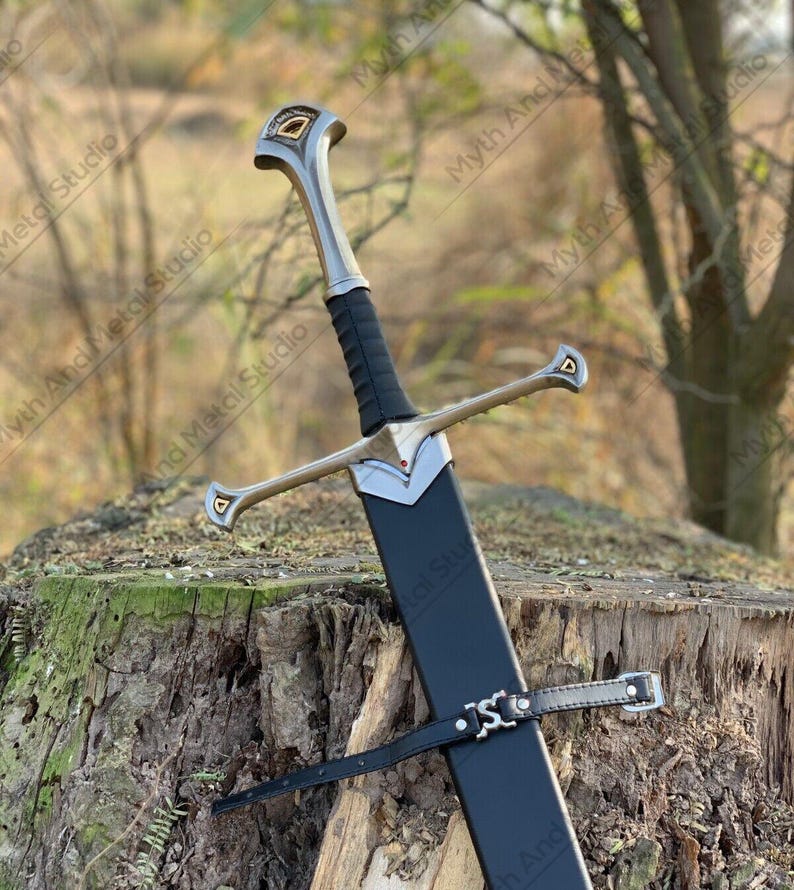 May include: A long, silver and black sword with a black sheath. The sword has a silver crossguard and a black handle. The sheath is secured with a black leather strap. The sword is resting on a tree stump.