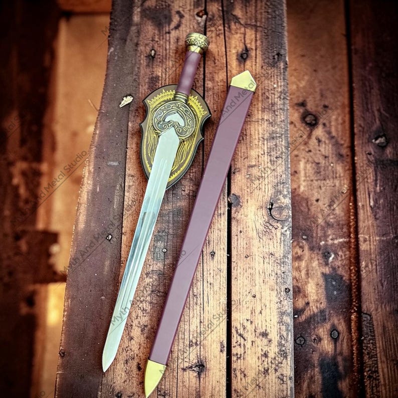 May include: A decorative sword with a silver blade, a burgundy handle, and a gold-colored hilt. The sword rests on a wooden surface next to a burgundy sheath with gold accents and a decorative shield.