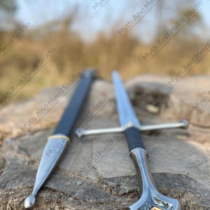 May include: A silver sword with a black handle and crossguard, alongside a black sheath with gold detailing. The sword features a pointed blade and an ornate handle. The background is blurred, with the text "Myth And Metal Studio" visible.