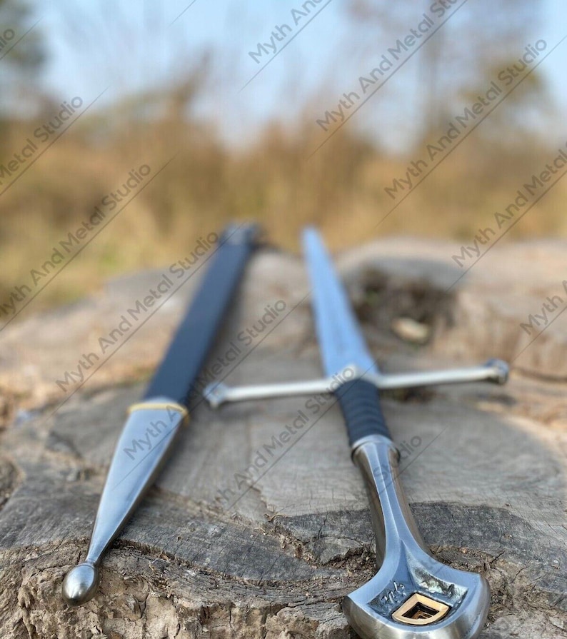 May include: A silver sword with a black handle and crossguard, alongside a black sheath with gold detailing. The sword features a pointed blade and an ornate handle. The background is blurred, with the text "Myth And Metal Studio" visible.