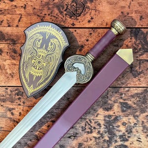 May include: A decorative sword with a silver blade, burgundy handle, and gold accents. The sword is paired with a matching burgundy sheath and a shield featuring a gold emblem of two horses. The shield is brown with gold trim.