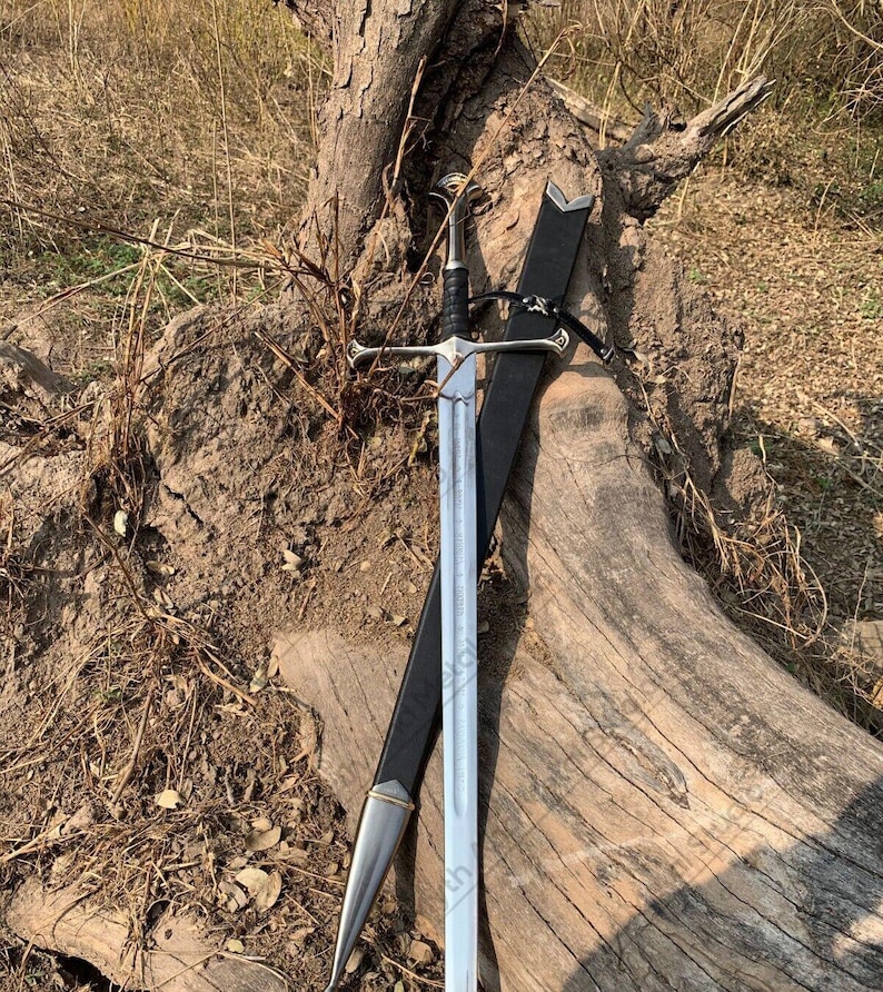 May include: A silver and black sword with a cross-shaped hilt and a leather sheath. The sword is leaning against a weathered tree trunk, with the blade partially drawn. The handle is black, and the blade is silver.