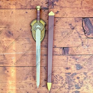 May include: A decorative sword with a silver blade, brown handle, and gold accents. The sword is displayed with a matching brown sheath and a decorative shield-shaped plaque. The plaque and sheath have gold-colored details.
