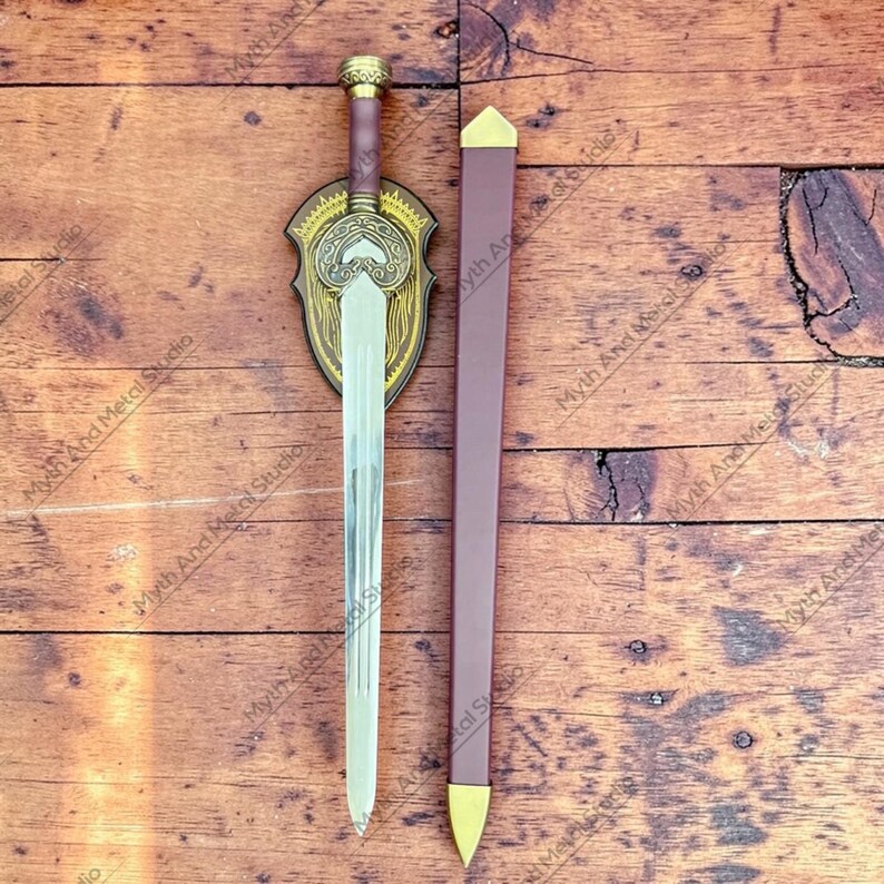 May include: A decorative sword with a silver blade, brown handle, and gold accents. The sword is displayed with a matching brown sheath and a decorative shield-shaped plaque. The plaque and sheath have gold-colored details.