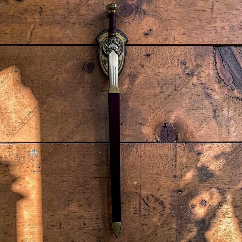 May include: A decorative sword with a gold and silver blade, a burgundy handle, and a gold-colored pommel. The sword is mounted on a shield-shaped plaque with intricate gold detailing. The background is a wooden surface.