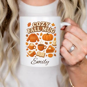 Puede incluir: Taza de cerámica blanca con el texto "COZY FALL MUG" y un diseño de temática otoñal. El diseño incluye una calabaza, un suéter, tazas de café, tarta y hojas. El nombre "Emily" está impreso debajo.