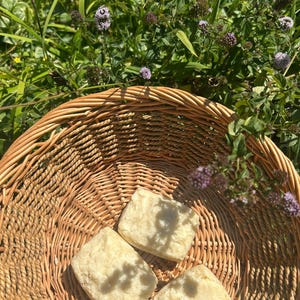 May include: A woven wicker basket holds three square, light-colored soaps. The basket is set amidst green foliage and purple wildflowers, suggesting a natural setting. The soaps appear handmade, with a slightly textured surface.