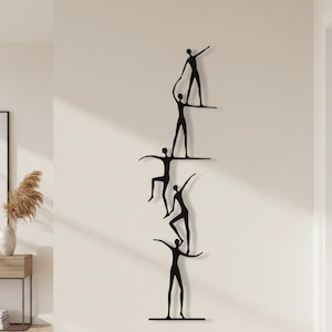 Könnte beinhalten: Eine vertikale Wandskulptur aus schwarzem Metall, die fünf stilisierte menschliche Figuren in einer ausbalancierten Formation zeigt. Jede Figur hat dünne, längliche Gliedmaßen, die ein durchgehendes, miteinander verbundenes Design an der Wand bilden.