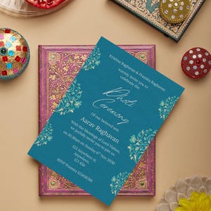 May include: A teal invitation card with gold floral accents, announcing a ceremony. The card is placed on a purple and gold decorated book. Other decorative items include a pomegranate and a gold and red jeweled ornament.