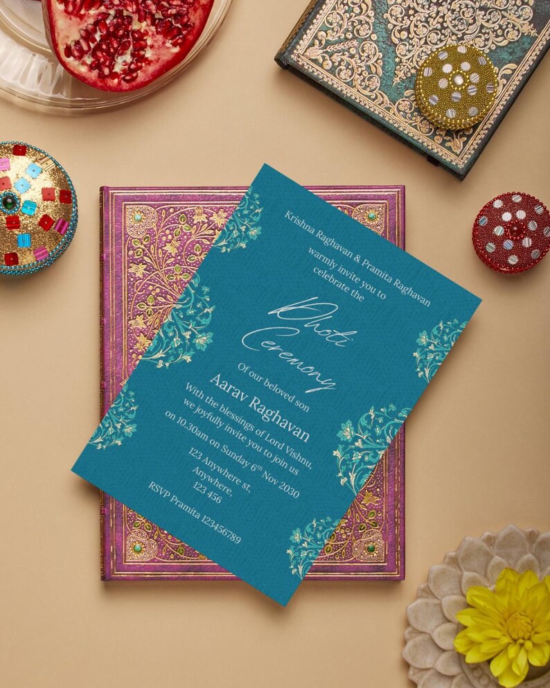 May include: A teal invitation card with gold floral accents, announcing a ceremony. The card is placed on a purple and gold decorated book. Other decorative items include a pomegranate and a gold and red jeweled ornament.