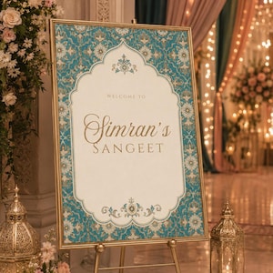 May include: A rectangular welcome sign in a gold frame on a tripod stand. The sign features a cream-colored center with the text "WELCOME TO Simran's SANGEET" in gold lettering, surrounded by a teal and gold floral pattern border with scalloped edges.