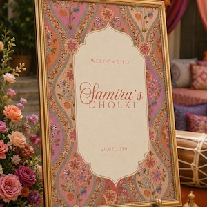 Op de afbeelding: Een rechthoekig bord in een gouden lijst op een metalen ezel. Het bord heeft een crèmekleurig midden met de tekst "WELCOME TO Samira's DHOLKI" en "29.07.2030". De rand heeft een roze, oranje en paars bloemenpatroon met fijne details.