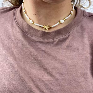 May include: A white beaded necklace with gold accents and a gold starfish pendant. The necklace also features white seashells. The necklace is worn on a person wearing a mauve t-shirt.