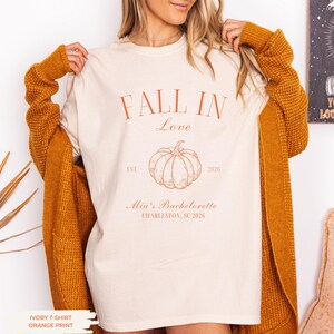 May include: An ivory t-shirt featuring orange text that reads FALL IN Love, EST. 2026, Mia's Bachelorette, and CHARLESTON, SC 2026, with a central line-art pumpkin illustration. The shirt is worn under a textured mustard yellow long-sleeve cardigan.