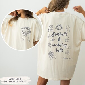 Puede incluir: Camiseta color marfil con estampado azul denim. La parte delantera presenta un diseño de concha con el texto "Megan's Bachelorette". La parte trasera muestra "Seashells & Wedding Bells" con ilustraciones de copas de champán, campanas y anillos. También está impresa la ubicación "Miami, FL".