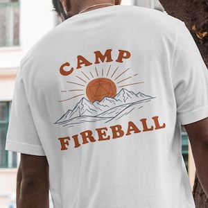 K&ouml;nnte beinhalten: Wei&szlig;es T-Shirt mit einem orangefarbenen Aufdruck auf dem R&uuml;cken. Das Design enth&auml;lt die Worte "CAMP FIREBALL", eine Sonne, Berge und eine geometrische Form. Das T-Shirt ist kurz&auml;rmelig.