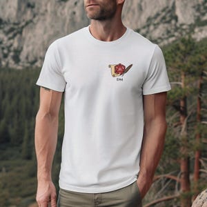Puede incluir: Camiseta blanca con un pequeño gráfico en el pecho. El gráfico presenta un dado rojo de veinte caras, un pergamino y una pluma, con las letras "DM" debajo. La camiseta está hecha de un material suave.