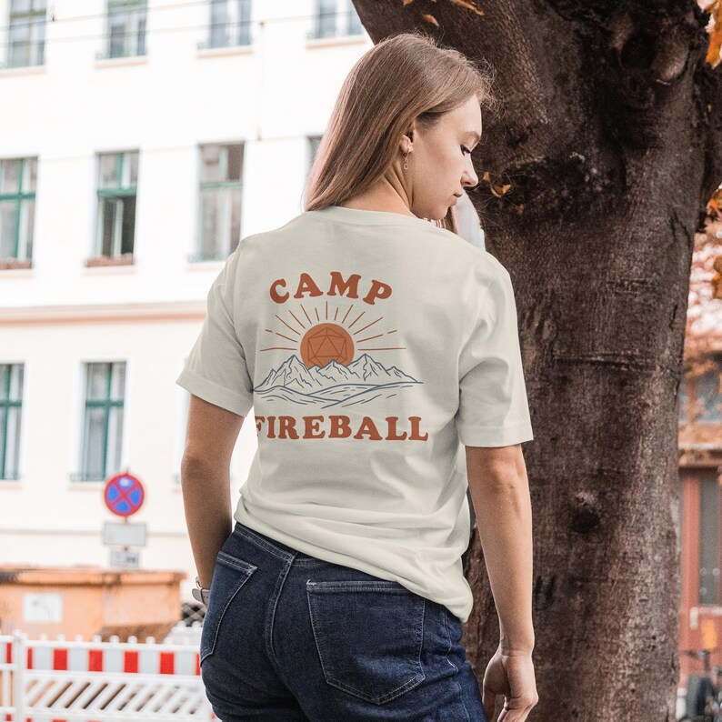 K&ouml;nnte beinhalten: Cremefarbenes T-Shirt mit den Worten "CAMP FIREBALL" in orangefarbenen Buchstaben. Das Design zeigt eine Sonne, Berge und eine geometrische Form. Das Shirt ist kurz&auml;rmlig und hat einen Rundhalsausschnitt.