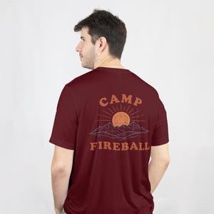 K&ouml;nnte beinhalten: Burgunderfarbenes T-Shirt mit einem grafischen Design auf dem R&uuml;cken. Das Design zeigt die Worte "CAMP FIREBALL" in Orange, mit einer Sonne und einer Bergillustration. Das T-Shirt ist einfarbig und hat kurze &Auml;rmel.
